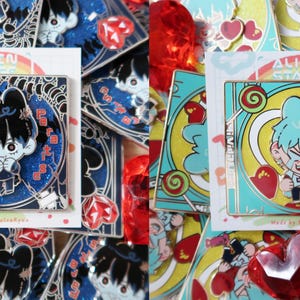 Peut inclure: Épingles en émail représentant des personnages de style anime avec des motifs de cœurs et de sucettes. Les épingles ont un design carré avec le texte "ALIEN STAGE" et du texte japonais. Les épingles sont sur une carte blanche avec un fond bleu et scintillant.