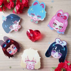 May include: A collection of colorful anime-style character acrylic pins and a red heart-shaped gem. The pins feature various hairstyles and bow accents in shades of blue, pink, brown, and blonde. Red flowers and glittery stars are scattered around the pins.