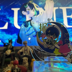 Puede incluir: Un soporte acrílico transparente con un personaje de dibujos animados en una luna creciente con acentos coloridos. El fondo muestra una imagen azul con la palabra "LUNE". Otras figuras son visibles en el fondo.