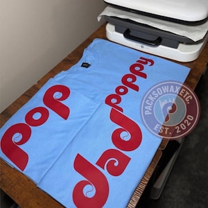 May include: Light blue t-shirts with the word "pop poppy" in red script. The shirts are on a wooden surface with a heat press in the background. A circular sticker with the text "PACKSOWAXETC. EST. 2020" is also visible.