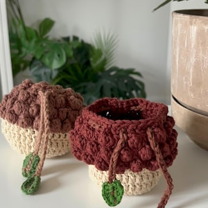 May include: Two small, round, crocheted pouches shaped like acorns. Each pouch features a textured, dark brown cap made with bobble stitches and a smooth, cream-coloured base. Brown drawstring cords hang from the sides, each ending in a small green leaf.
