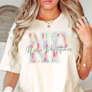 May include: A white t-shirt with a colorful "NP" design that says "Nurse Practitioner" underneath. The shirt is being worn by a person with long blonde hair.