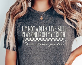 May include: A dark gray t-shirt with a white graphic that reads "I'm not a detective, but I play one from my couch. True crime junkie." The graphic includes a black and white checkered pattern.