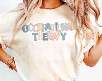 May include: A white t-shirt with the text "OCCUPATIONAL THERAPY" in colorful letters. The letters are outlined in black and have a slightly distressed look. The shirt is being worn by a person with long blonde hair and is paired with blue jeans.