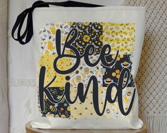 May include: A white canvas tote bag with black straps and a patchwork design featuring bees, honeycombs, and the text "Bee Kind".