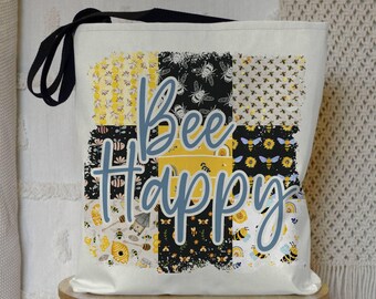 May include: A white canvas tote bag with black straps and a patchwork design featuring bees, honeycombs, and sunflowers. The text "Bee Happy" is printed in blue on the bag.