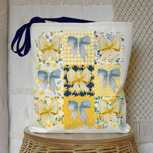 May include: A white canvas tote bag with navy blue handles. The bag features a patchwork design with yellow and blue bows on a white background.