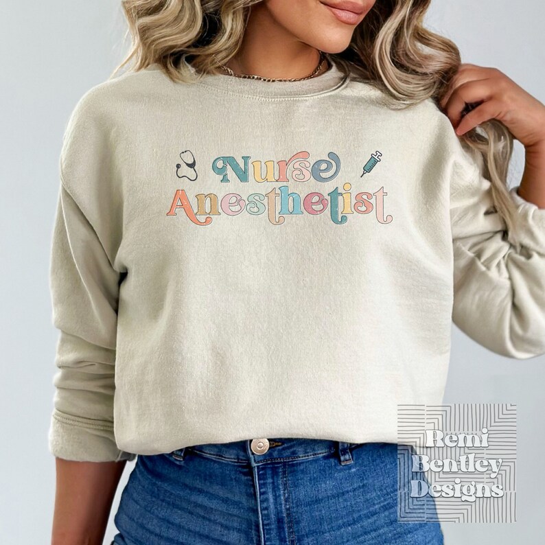May include: A light beige sweatshirt with the text "Nurse Anesthetist" in colorful letters. The text is surrounded by a stethoscope and a syringe.