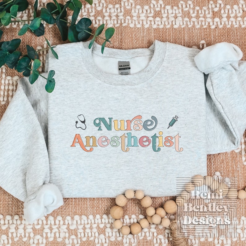 May include: A gray sweatshirt with the text "Nurse Anesthetist" in colorful retro-style lettering. A stethoscope and syringe are included in the design.