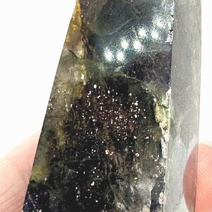 May include: A dark, faceted crystal with a mix of green, black, and brown hues. The crystal has a polished surface with sparkling inclusions, and a lighter green tip. The overall shape is a rectangular prism with a slanted top.