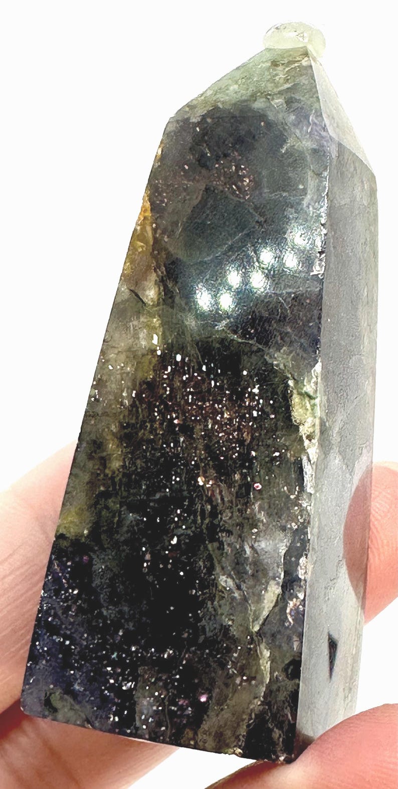 May include: A dark, faceted crystal with a mix of green, black, and brown hues. The crystal has a polished surface with sparkling inclusions, and a lighter green tip. The overall shape is a rectangular prism with a slanted top.