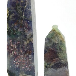 May include: Two polished gemstone obelisks. The larger obelisk is a deep purple with green and pink inclusions. The smaller obelisk is green and brown with a rounded top. Both are set against a white background.