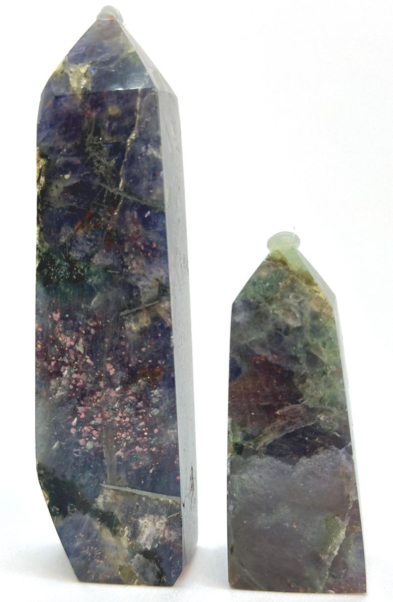 May include: Two polished gemstone obelisks. The larger obelisk is a deep purple with green and pink inclusions. The smaller obelisk is green and brown with a rounded top. Both are set against a white background.