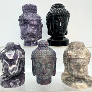 May include: Five carved stone Buddha head sculptures in various colors. The sculptures include purple, black, and gray tones. The Buddha heads are displayed on a clear shelf against a white background. The sculptures are detailed and have a serene expression.