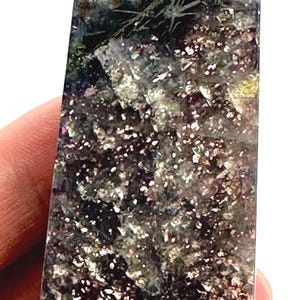 May include: A rectangular crystal obelisk with a polished surface. The stone features a mix of colors, including dark purple, silver, and green, with sparkling inclusions. The obelisk is held in a hand.