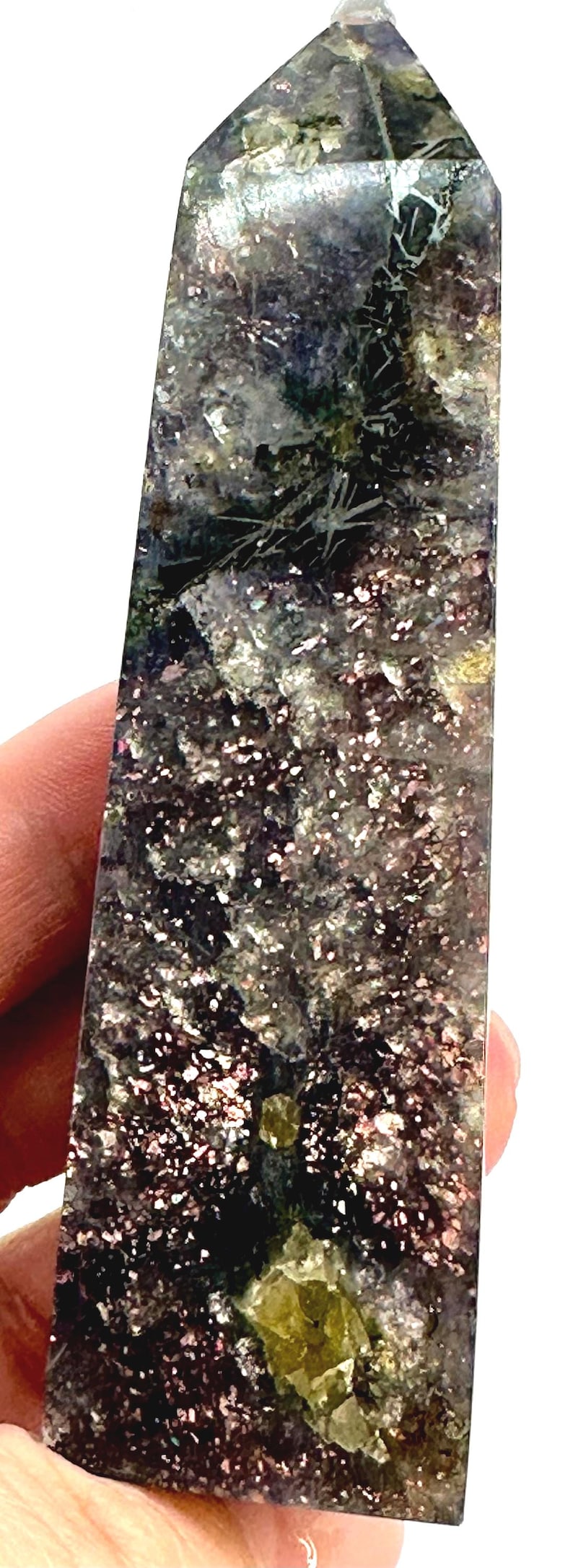 May include: A rectangular crystal obelisk with a polished surface. The stone features a mix of colors, including dark purple, silver, and green, with sparkling inclusions. The obelisk is held in a hand.
