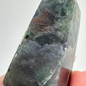 May include: A polished, multi-colored crystal point. The crystal features shades of green, purple, and brown. The top of the crystal has a small, clear, rounded bead. The crystal is held by a person's fingers.