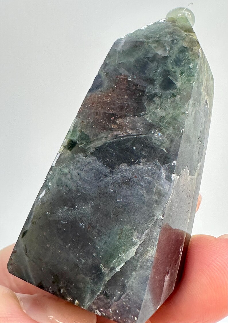 May include: A polished, multi-colored crystal point. The crystal features shades of green, purple, and brown. The top of the crystal has a small, clear, rounded bead. The crystal is held by a person's fingers.