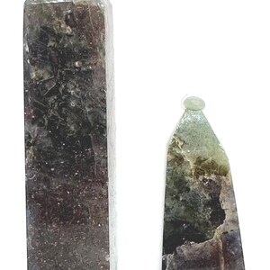 May include: Two natural stone obelisks. The taller one is rectangular with a pointed top, and the shorter one is triangular. Both have a mix of dark green, brown, and gray tones. The obelisks are set against a white background.