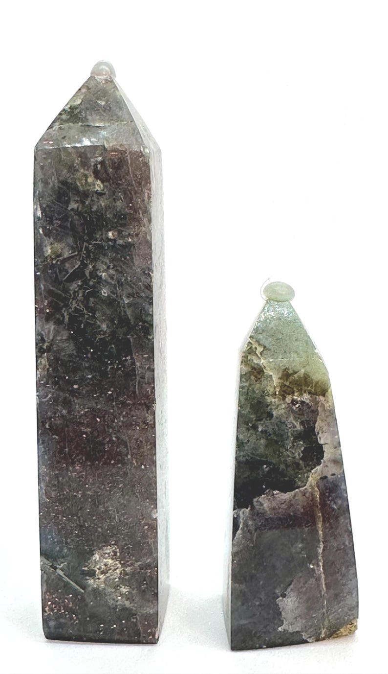 May include: Two natural stone obelisks. The taller one is rectangular with a pointed top, and the shorter one is triangular. Both have a mix of dark green, brown, and gray tones. The obelisks are set against a white background.