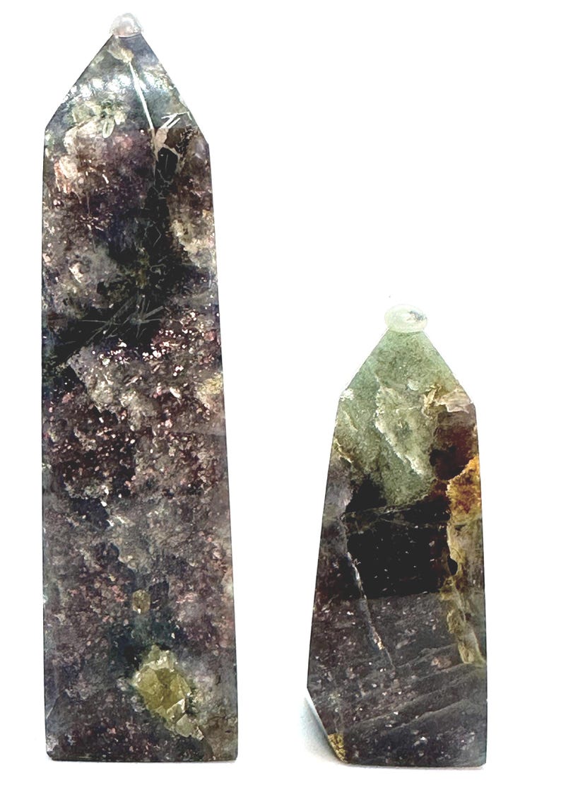 May include: Two polished crystal obelisks. The larger obelisk is a dark purple and gray color with flecks of gold and green. The smaller obelisk is green, brown, and purple. Both have a clear crystal tip.