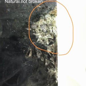 May include: Close-up of a dark, textured stone with the text "Natural not broken." The stone has a rough, uneven surface with areas of green and white. The edge of the stone is highlighted with an orange circle.