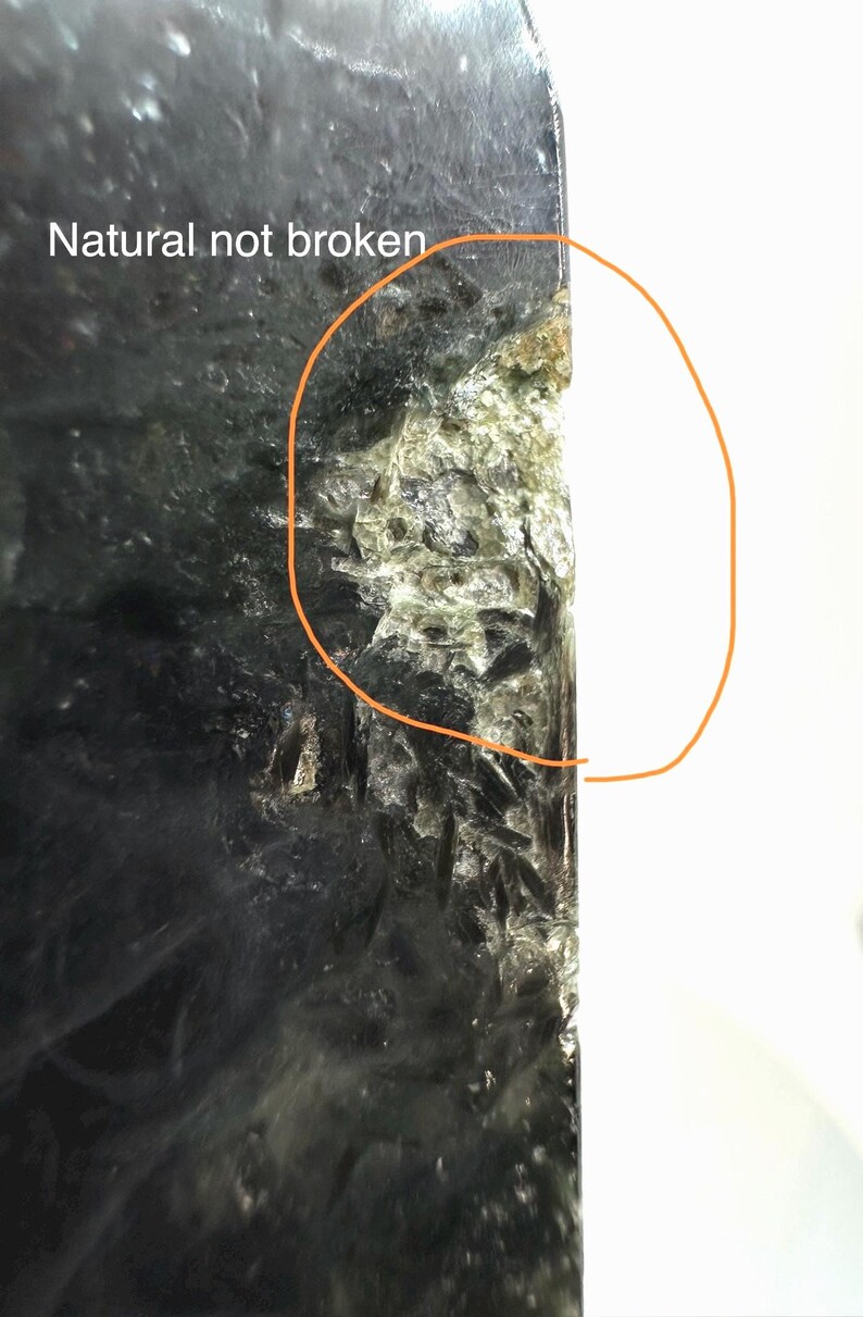 May include: Close-up of a dark, textured stone with the text "Natural not broken." The stone has a rough, uneven surface with areas of green and white. The edge of the stone is highlighted with an orange circle.