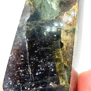 May include: A polished crystal point with a mix of colors, including green, black, and gold. The crystal has a faceted shape and a translucent appearance. The surface has a sparkling effect, and the crystal is held by a person's fingers.