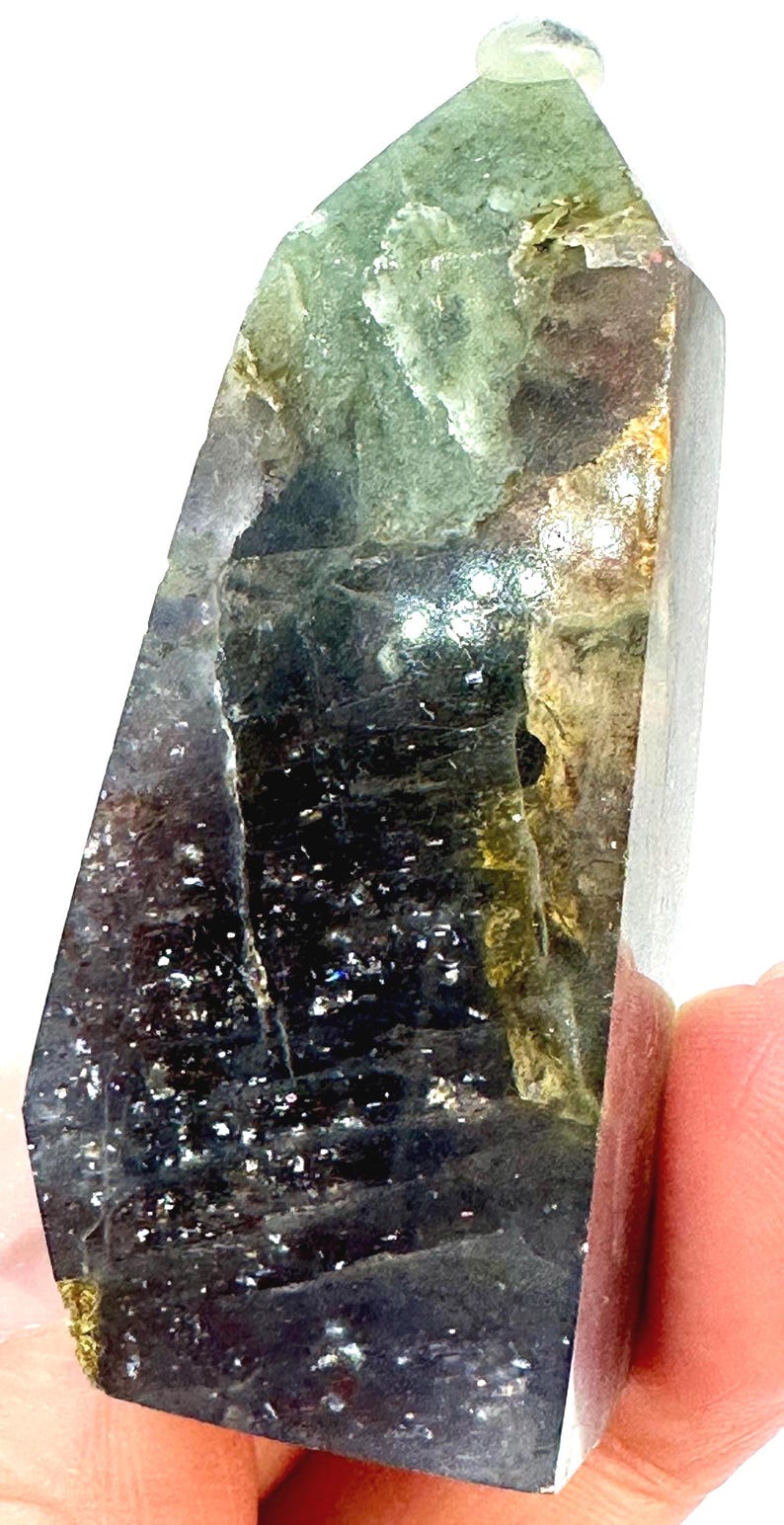 May include: A polished crystal point with a mix of colors, including green, black, and gold. The crystal has a faceted shape and a translucent appearance. The surface has a sparkling effect, and the crystal is held by a person's fingers.
