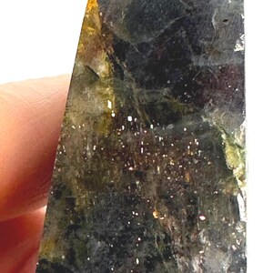 May include: A polished, triangular-shaped gemstone obelisk. The stone features a dark, mottled appearance with shades of green, black, and gold. Tiny, sparkling inclusions are scattered throughout the surface, adding a glittery effect.