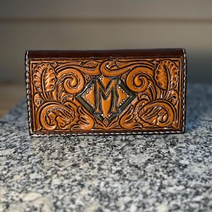 May include: A brown leather wallet with intricate floral and scrollwork carving. The center features a diamond-shaped design with the letter "M". The edges are stitched with white thread, and the wallet has a dark brown border.