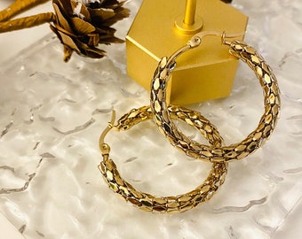 May include: A pair of gold hoop earrings with a textured, chain-like design. The earrings are displayed on a clear, textured surface.