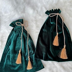 May include: Two green velvet drawstring bags with gold tassels. The bags are on a white fluffy surface.