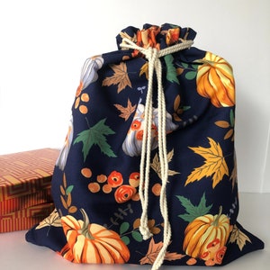 May include: A navy blue drawstring bag with a fall-themed pattern. The fabric features orange pumpkins, fall leaves, and floral accents. A cream-colored rope cinches the top. A decorative box is partially visible in the background.