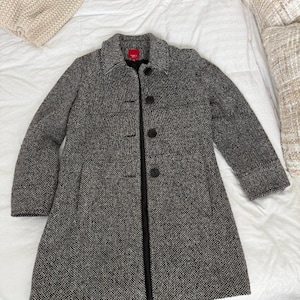 May include: A black and white tweed coat featuring a collared neckline, long sleeves, and a front button closure with four visible dark buttons. The interior label is red with white text. The coat has a textured, woven fabric construction.