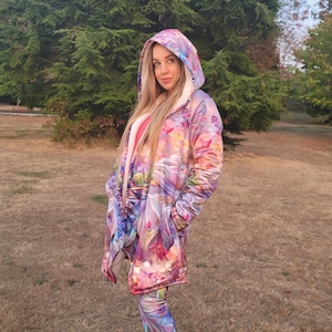 May include: A long-sleeved hooded jacket with a colorful floral and butterfly print. The jacket has a white sherpa lining and is worn over leggings with a similar print. The person is standing in a grassy field.