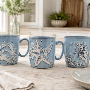 May include: Three light blue ceramic mugs featuring raised white relief designs of a crab, a starfish, and a seahorse. Each mug includes a handle and decorative coral accents, showcasing a coastal theme with a smooth, glazed finish throughout.