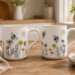 May include: Two white ceramic mugs with yellow interiors, each featuring black line-art illustrations of wildflowers and bumblebees. A circular stamp on the upper side of each mug reads Original Hand Painted. The mugs have a sturdy handle and base.