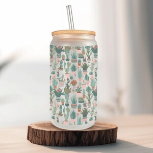 May include: A frosted glass can-shaped tumbler with a bamboo lid and a metal straw. The tumbler is decorated with a repeating pattern of various potted cacti and succulents in shades of green, pink, and beige. The tumbler sits on a small, round wooden coaster.