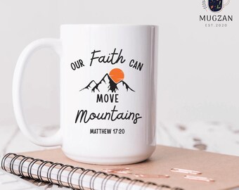May include: White ceramic mug featuring the phrase "Our Faith Can Move Mountains" in black script, with a mountain and sun graphic. The text "Matthew 17:20" is below. The mug rests on a spiral-bound notebook.