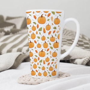 May include: A white latte mug with a repeating pattern of orange pumpkins and autumn leaves. The mug has a large handle and is sitting on a beige coaster. The background includes a white surface and a patterned blanket.