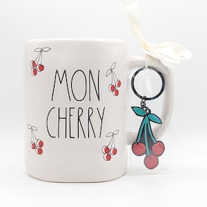 May include: White ceramic mug with the words "MON CHERRY" and cherry illustrations. A cherry-shaped keychain with red cherries and teal leaves hangs from the mug's handle. The mug is a gift set.