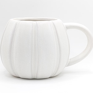 Könnte beinhalten: Eine weiße Keramik-Tasse in Form eines Kürbisses. Die Tasse hat vertikale Rillen und einen gebogenen Henkel. Die Tasse ist einfarbig weiß und steht auf einer weißen Oberfläche.