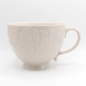May include: A beige ceramic teacup with a floral embossed design. The cup has a large handle and sits on a small pedestal base. The floral pattern includes roses and leaves.