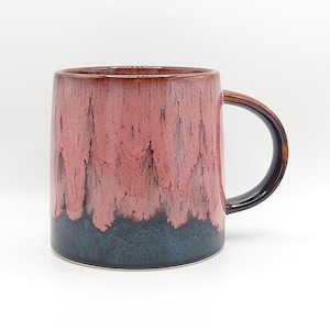 May include: A ceramic mug featuring a reactive glaze with a dusty rose pink upper section that drips down into a dark charcoal gray base. The mug has a smooth, cylindrical body and a curved handle that matches the dark gray color of the base.