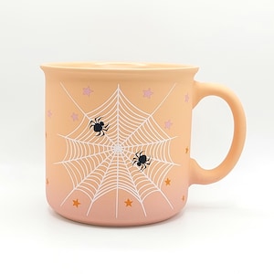 May include: A peach-colored ceramic mug featuring a white spiderweb graphic, two black spiders, and small scattered stars in shades of pink and orange. The mug has a smooth, matte finish and a curved handle on the right side of the cylindrical body.