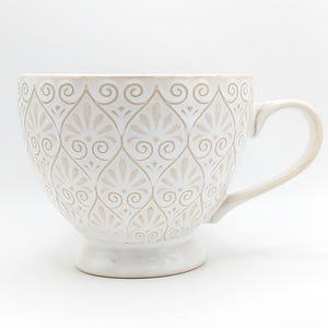 May include: White ceramic teacup with a beige floral pattern and a handle. The teacup has a wide bowl shape and sits on a small pedestal base. The design is symmetrical and covers the entire surface of the cup.