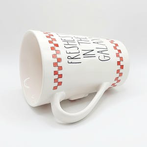 May include: White ceramic mug with a curved handle. The mug features a red and white checkered pattern around the rim and the words "Freshest in the Galaxy" in black lettering. The mug is angled, showing the interior.