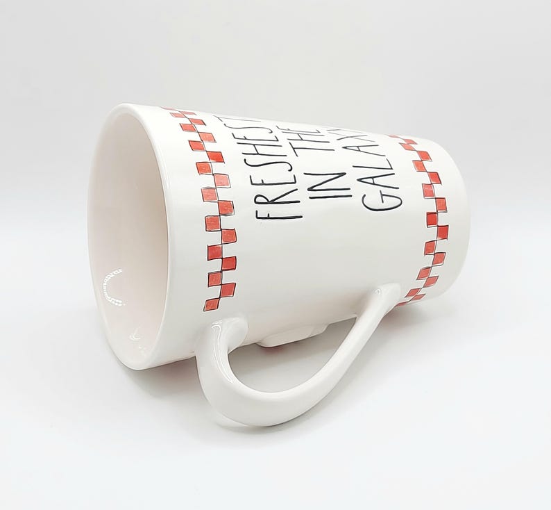 May include: White ceramic mug with a curved handle. The mug features a red and white checkered pattern around the rim and the words "Freshest in the Galaxy" in black lettering. The mug is angled, showing the interior.
