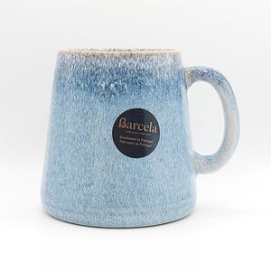 May include: A blue ceramic mug with a rounded handle. The mug has a light blue glaze with darker blue and brown accents near the rim. A black sticker on the mug reads "Barcela, Handmade in Portugal."